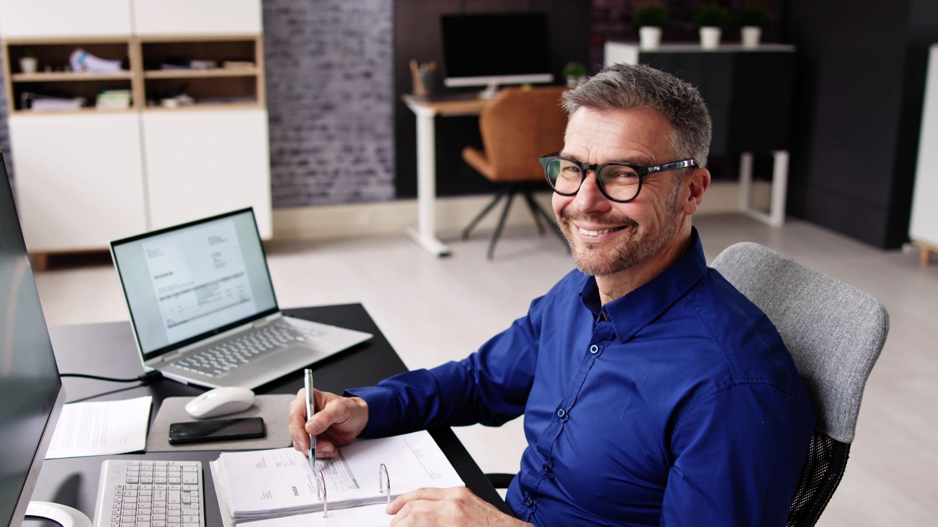 Senior finance professional reviewing invoices and accounting records at his desk in a corporate office.